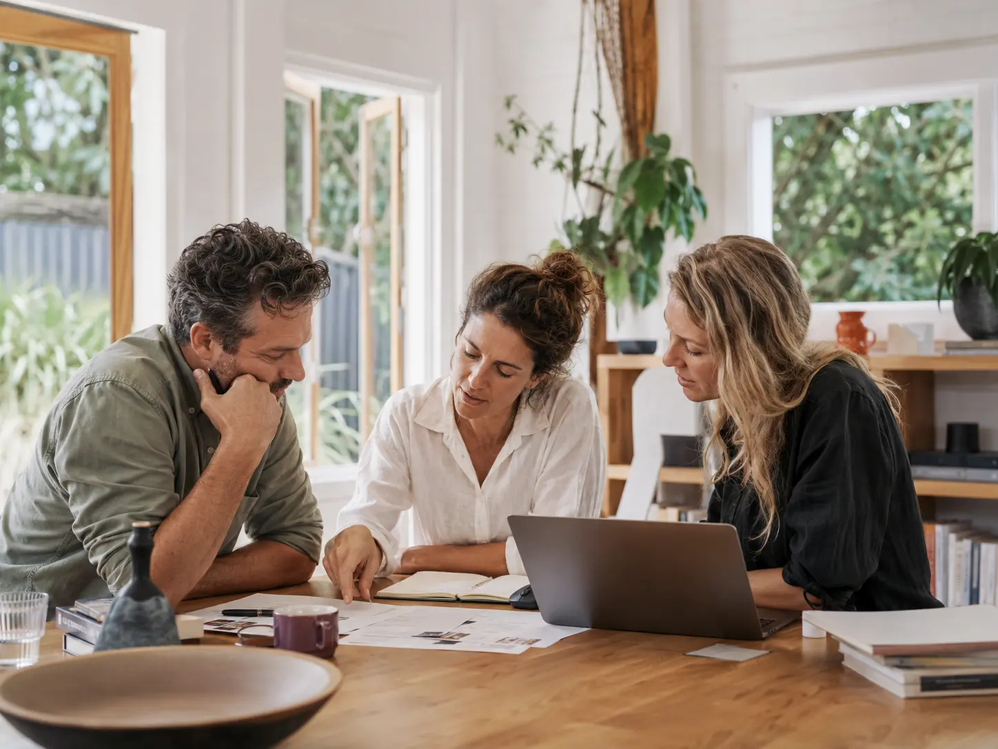 Three people in a bright studio discussing business work around a timber table.
